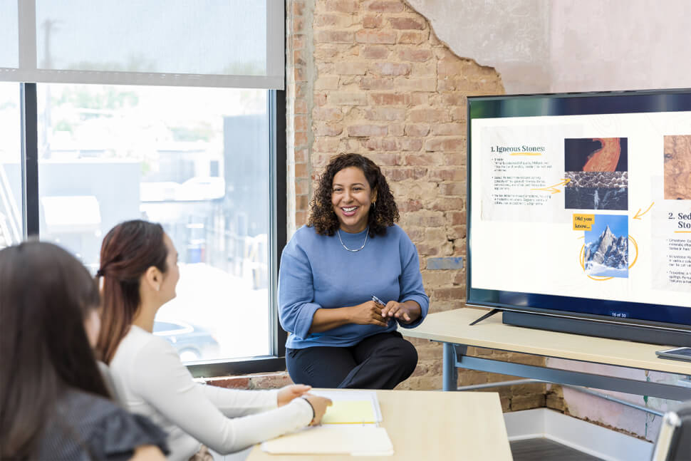 Teacher smiling sitting down showing a images on a screen while students sit down